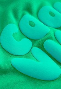 Zöld pamutszövet, amelyen nagy, kiemelkedő, matt türkizkék betűk láthatók, lekerekített formákkal és puha textúrával, amelyek merész, grafikus mintát alkotnak.