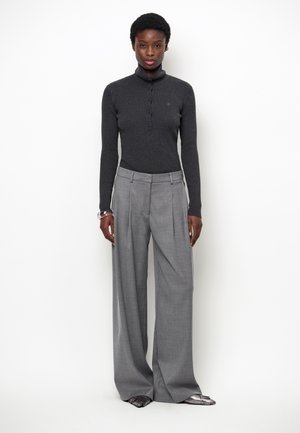 Vrouw met kort zwart haar die een donkere geribbelde coltrui en wijde grijze broek draagt, staand tegen een egale witte achtergrond.