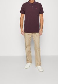 Polo tričko s krátkým rukávem v barvě burgundy, se dvěma knoflíky a malým červeným logem, spárované s béžovými kalhotami a bílými teniskami. Jednoduché pozadí.