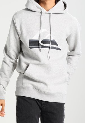 Homme portant un sweat à capuche gris clair avec un logo montagne abstrait noir et blanc, associé à un pantalon noir, sur un fond uni.