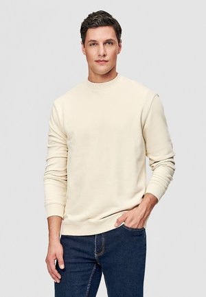 Mężczyzna w kremowej, długorękawowej bluzie i ciemnoniebieskich dżinsach, stojący z jedną ręką w kieszeni na jednolitym tle.