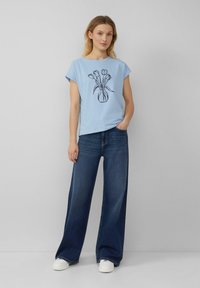 Hellblaues T-Shirt mit kurzen Ärmeln und schwarzem grafischen Blumenvasen-Motiv, kombiniert mit weit geschnittenen dunklen Jeans und weißen Turnschuhen. Einfaches Design und lässige Passform.