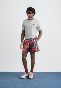 Γκρι κοντομάνικη μπλούζα Nike, πολύχρωμο σορτς με αφηρημένο μοτίβο, κόκκινες κάλτσες και γκρι αθλητικά παπούτσια, στέκεται μπροστά σε απλό φόντο.