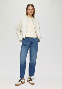 Jasno beżowa kurtka bomberka, kremowy sweter, niebieskie dżinsy i wielokolorowe trampki. Posiada zamek błyskawiczny i dwie boczne kieszenie.