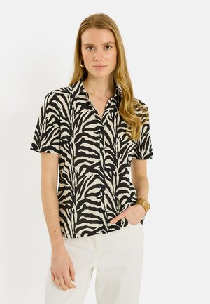 Femme aux longs cheveux blonds portant une chemise à manches courtes boutonnée à rayures zébrées noires et blanches et un pantalon blanc, debout devant un fond uni.