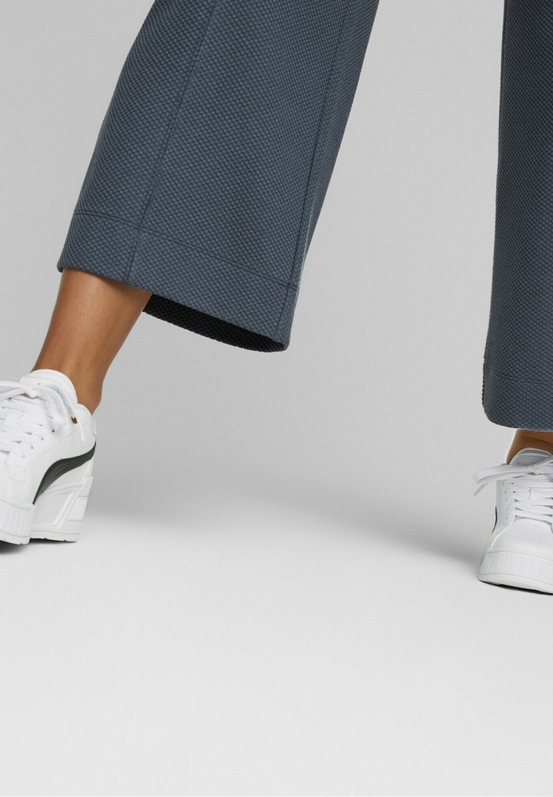 Jambes inférieures portant un pantalon large texturé foncé associé à des baskets blanches avec des bandes noires, sur un fond clair uni.