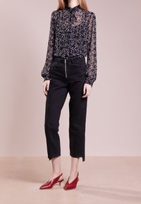 Femme portant un chemisier noir transparent à étoiles blanches, un pantalon noir court taille haute et des escarpins rouges à talons bas et bride arrière, debout.