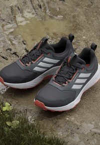 Chaussures de trail running Adidas Terrex noires et grises avec des bandes blanches et des touches orange reposant sur un sol boueux près de l'herbe.