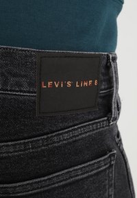 Jean en denim noir porté avec un haut bleu sarcelle, affichant une étiquette noire avec un texte rouge indiquant "LEVI'S LINE 8" sur la ceinture.