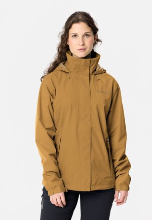 Frau mit langen lockigen Haaren, die eine senfbraune Jacke mit hohem Kragen und schwarze Hose trägt, steht vor einem einfarbigen hellen Hintergrund.