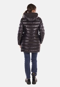 Giubbotto a vento nero con cappuccio, caratterizzato da un design a righe orizzontali e una texture lucida, abbinato a jeans scuri e stivali neri imponenti.