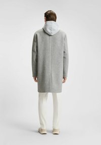 Cappotto di lana grigio con cappuccio grigio chiaro, taglio dritto e maniche lunghe. Indossato sopra pantaloni bianchi, presenta un design pulito e una texture morbida.