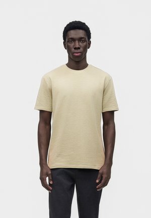 Man die recht vooruit kijkt, draagt een beige gestructureerd kort shirt en zwarte broek tegen een effen witte achtergrond.