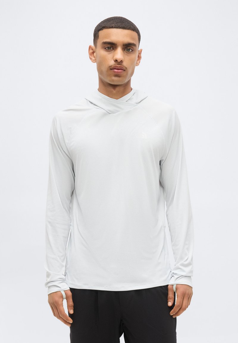 Jeune homme portant un sweat à capuche gris clair uni à manches longues avec des trous pour les pouces, debout devant un fond gris clair.