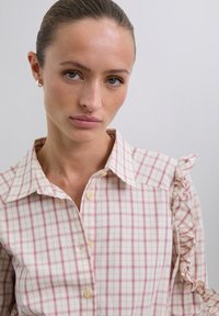 Blusa a cuadros en tejido suave con patrón de cuadrícula en rosa y blanco. Presenta un cuello clásico y detalle con volantes en el hombro.