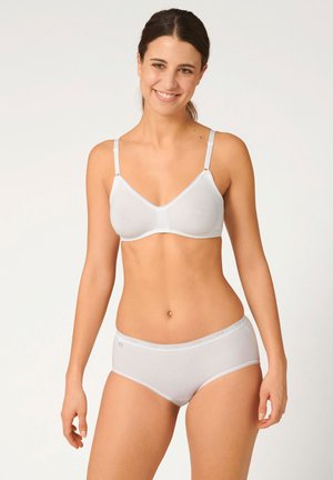 Donna con capelli scuri che indossa un set coordinato di reggiseno e slip bianchi, sorridente e in piedi davanti a uno sfondo chiaro uniforme.