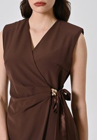 Braunes ärmelloses Wickelkleid aus glatter Stoffqualität, mit V-Ausschnitt, seitlichem Schleifenakzent mit goldenem Hardware-Detail und seitlicher Taschenanbringung.