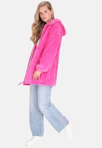 Růžový fleecový kabát s kapucí, zapínáním na zip a bočními kapsami, nošený přes světle modré džíny a bílé tenisky. Měkká textura.