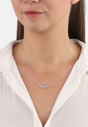 Donna che indossa una collana d'argento con piccoli ciondoli a forma di farfalla, abbinata a una camicia bianca con colletto.