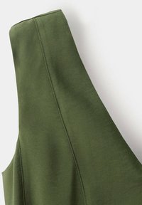 Gros plan d'un vêtement en tissu vert olive avec un col montant et des coutures verticales sur un fond clair uni.