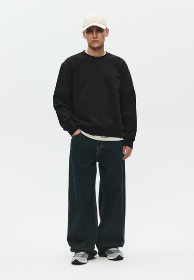 Joven con gorra beige, sudadera negra oversize, pantalones vaqueros anchos oscuros y zapatillas grises, de pie con una mano en el bolsillo.