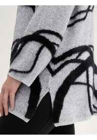 Pull en tricot gris avec des motifs abstraits noirs duveteux, présentant un ourlet côtelé et des fentes sur les côtés pour ajouter de la texture et des détails de design.