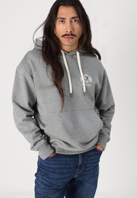Grauer Kapuzenpullover mit cremefarbenen Kordeln und Fronttasche, mit einem Logo auf der Brust. Getragen mit dunkelblauen Jeans.