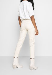 Pantalones vaqueros color crema con un corte ajustado, que cuentan con bolsillos traseros y costuras en contraste, combinados con una camisa blanca de manga corta y botas de color claro.
