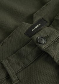 Nahaufnahme eines olivgrünen Stoffs mit strukturierten Oberfläche, versehen mit einem schwarzen Markenetikett und einem schwarzen Knopfdetail.