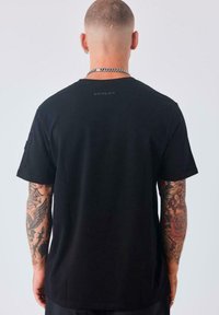 Hombre con cabello corto y tatuajes en ambos brazos, vestido con una camiseta negra y una cadena de plata, visto desde la espalda.