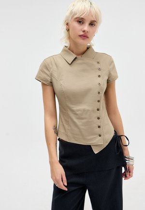 Jeune femme aux cheveux blonds courts porte un haut beige asymétrique boutonné et un pantalon noir à fines rayures, posant devant un fond uni.