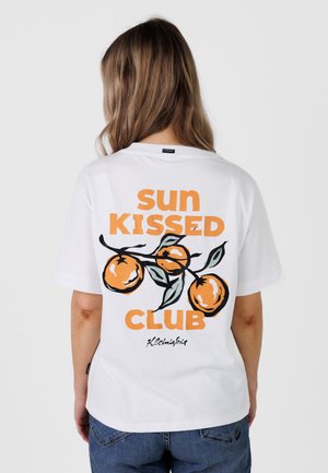 Kvinde med langt hår iført en hvid t-shirt med appelsinfrugt og teksten "sun kissed club" på ryggen, kombineret med blå jeans.
