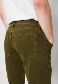 Olive green cotton pants with a smooth texture, featuring back pockets, subtle stitching, and a small embroidered logo detail.