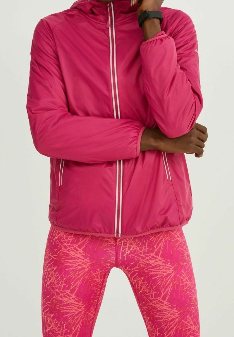 Personne portant une veste zippée rose vif et des leggings roses à motifs, les bras croisés et une montre connectée au poignet gauche.