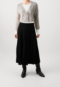 Světle šedý pletený cardigan s uvazováním vpředu, přes bílý dlouhý rukáv top. Černá plisovaná midi sukně a černé kotníčkové boty dokončují outfit.