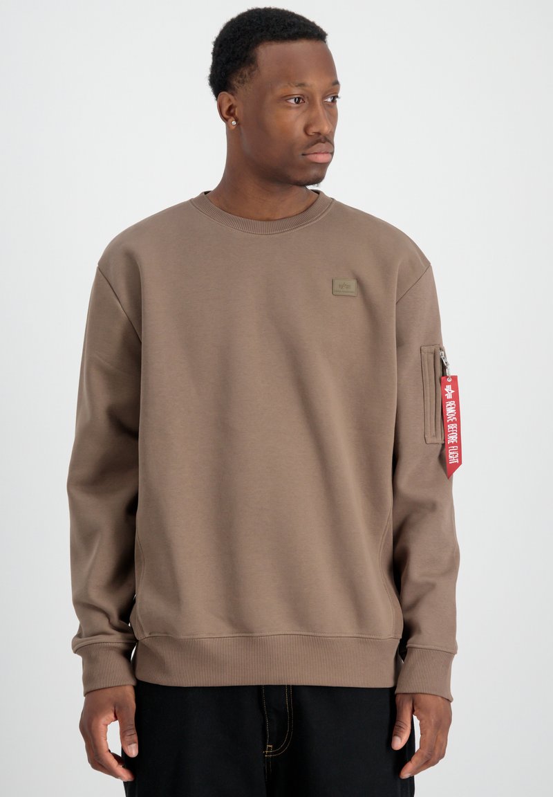 Felpa marrone realizzata in morbido materiale, con colletto e polsini a costine, una tasca laterale con zip e un patch con il marchio sul petto.