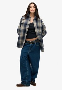 Flannel skjorte i blå og creme tern, åben over en sort tanktop. Løse blå jeans med et sort bælte. Brune slip-on sko.