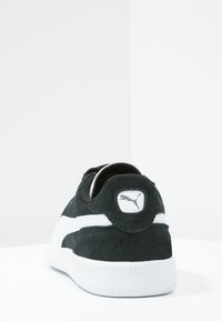 Schwarze Wildleder-Sneaker mit weißer Gummisohle, ausgestattet mit einer gepolsterten Ferse und einem Logopatch. Das Design umfasst weiße Streifen an den Seiten.