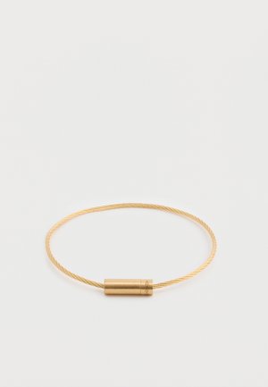 Kuldne käevõru, millel on keerdsellede disain ja silindriline lukk, millel on graveeritud aksessuaarid. Sile tekstuur metallikviimistlusega.