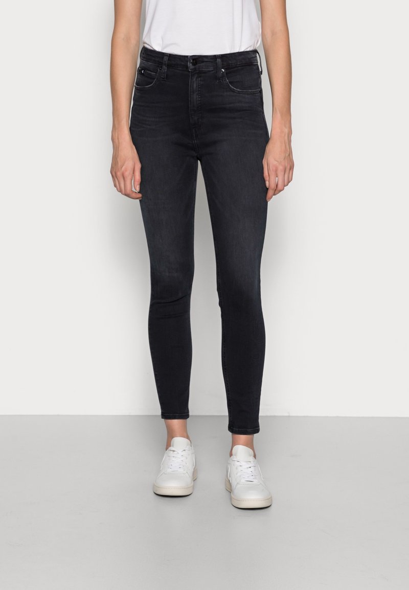 Jeans ajustados en denim oscuro, con una cintura alta, diseño de cinco bolsillos y detalles de costura sutiles. Combinados con zapatillas blancas.