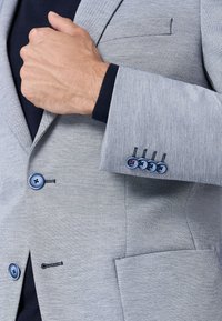 Blazer grigio intrecciato con vestibilità sartoriale, bottoni blu e una piccola tasca; tessuto strutturato con cuciture dettagliate sui polsini.