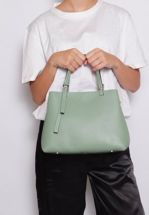 Helle grüne Lederhandtasche mit strukturiertem Design, doppelten kurzen Griffen, dezenten Hardware-Akzenten und texturierter Oberfläche, gehalten von einer Person.