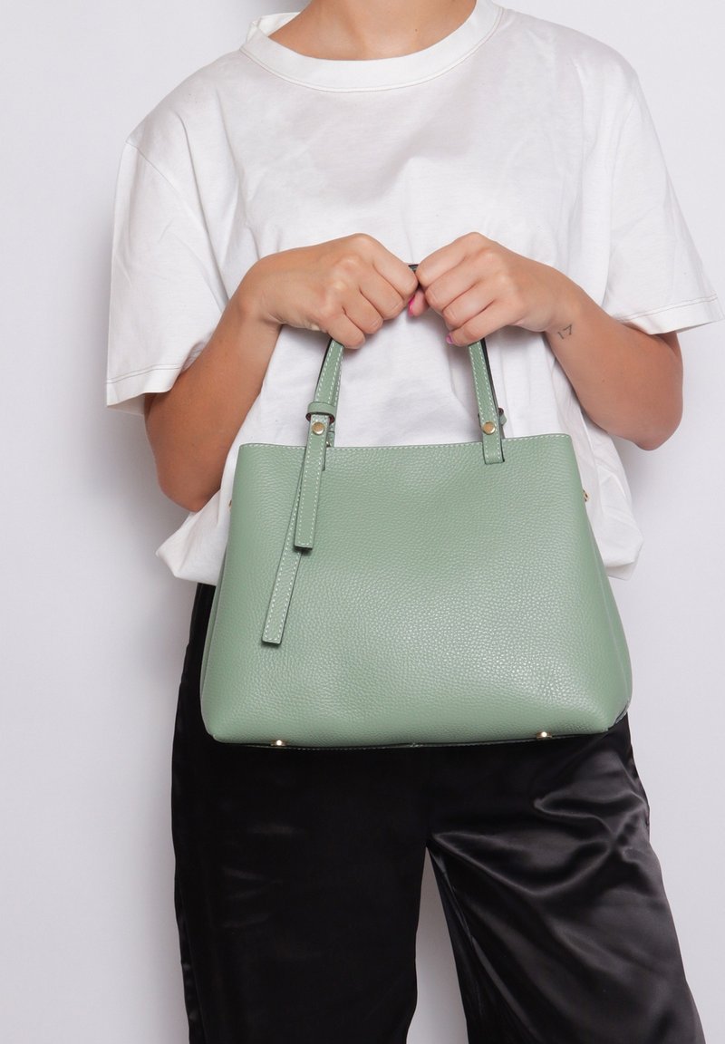 Lichtgroene leren handtas met gestructureerd ontwerp, dubbele korte handvatten, subtiele hardware-accenten en een textuurfinish, vastgehouden door een persoon.
