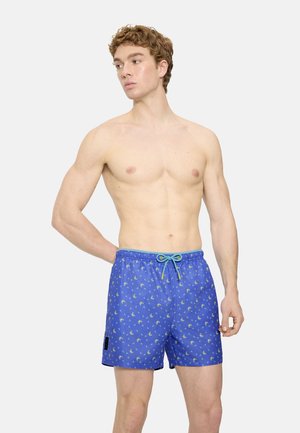 Joven sin camisa que lleva pantalones cortos de baño azules con un pequeño patrón floral, de pie contra un fondo claro y liso.