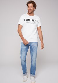 Camiseta blanca con texto negro "CAMP DAVID" y detalles adicionales de marca, combinada con jeans de color azul claro y zapatillas blancas.