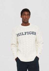 Cremiger, kabelstrukturierter Pullover mit gerippten Bündchen und Saum, ausgestattet mit einem auffälligen marineblauen "HILFIGER"-Logo über der Brust. Weiche Textur und lässige Passform.