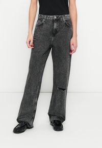 Fekete farmer széles szárú nadrág sérült térddel, hagyományos ötlovas zsebes kialakítással és fém kiegészítőkkel, fekete cipővel párosítva.