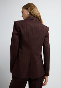 Mujer con cabello ondulado vistiendo un traje a rayas en color marrón oscuro, vista desde atrás contra un fondo claro y liso.