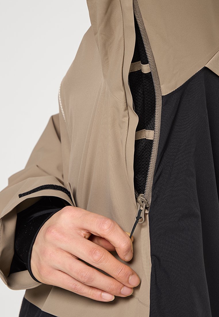 Beige und schwarze Jacke mit Reißverschluss, die ein strukturiertes Innenfutter, einen lockeren Schnitt und elastische Bündchen aufweist. Handgriffe am Reißverschluss ziehen für den Verschluss.