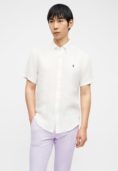 Άνδρας που φοράει λευκό πουκάμισο με κοντό μανίκι και μπλε λογότυπο, καθώς και ανοιχτόχρωμο μωβ παντελόνι, στέκεται με το ένα χέρι στην τσέπη.